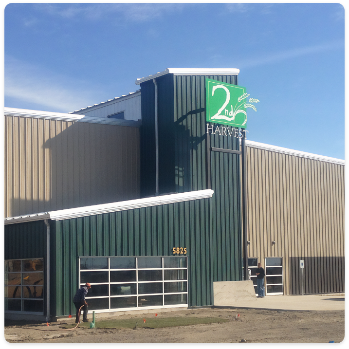 A green and beige building with a sign that reads "2nd Harvest" on a tall section of the exterior. The address number "5825" is displayed above a garage door. A person from a commercial painting service is kneeling near the base of the building. The sky above is clear and blue.