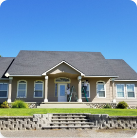 Single-story beige house with a dark gray roof, featuring a covered front porch with white columns and trimmed hedges. Two people on ladders, likely offering commercial painting services, are working near the front entrance. The sky is clear and blue.