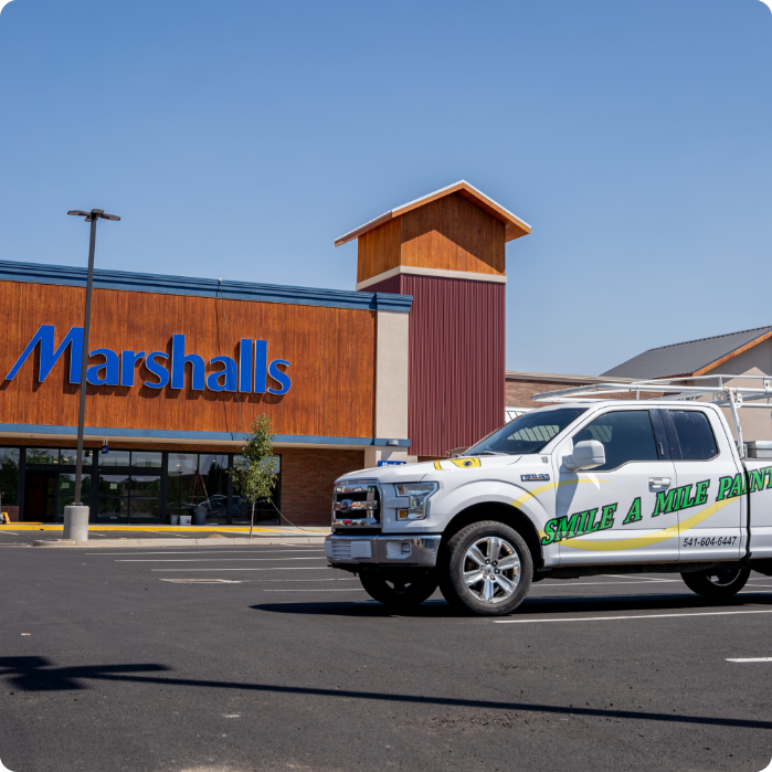 A "Marshalls" store is visible against a clear sky, with a parking lot in front. A white truck with green lettering that reads "SMILE A MILE PAINT – Commercial Painting Services" and a phone number is parked near the store.