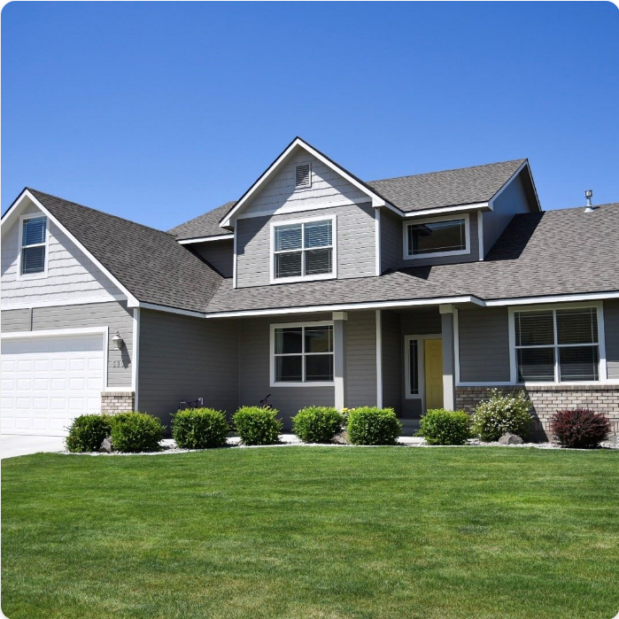 A modern two-story house with gray siding, white trim, and a dark shingled roof. The front features a garage on the left, a porch with a yellow door, two large front-facing windows, and a manicured lawn with trimmed bushes lining the walkway. Perfect for showcasing top-notch exterior painting services.