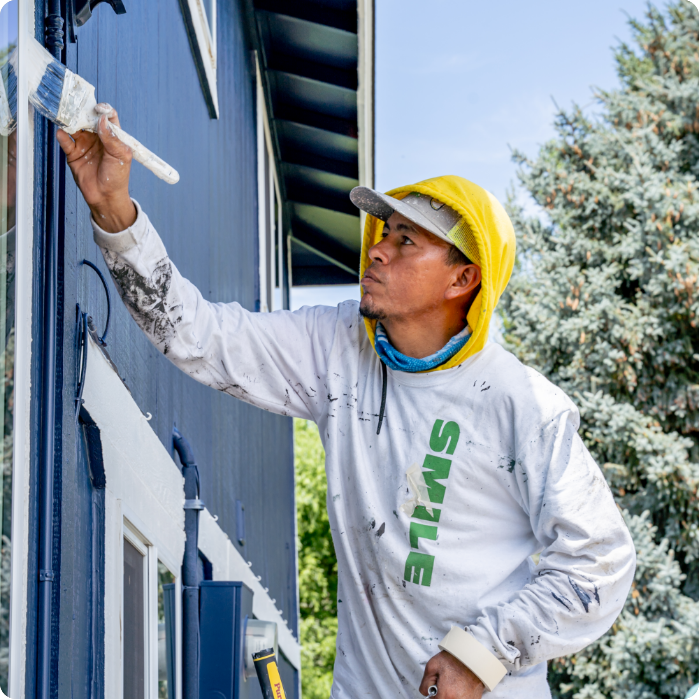 A man in a yellow hat and white hoodie, with "SMILE" written on it, is focused on painting the exterior of a house blue. Holding a paintbrush, he reaches up to paint near a window. The area around him is splattered with paint, and trees are visible in the background—showcasing his commercial painting skills.