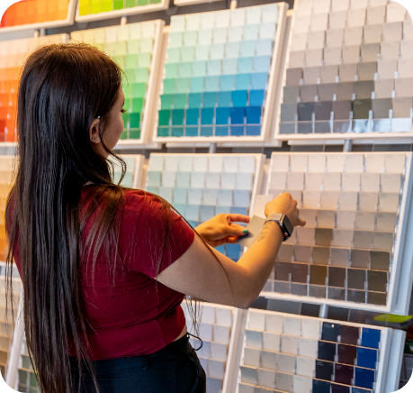 A person with long hair wearing a red shirt and black pants is selecting color swatches from a display wall filled with various shades arranged in rows. The wall includes a wide range of colors, perfect for anyone seeking inspiration for commercial painting services, covering both interior and exterior designs.