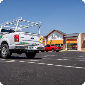 A white truck with a painting company’s logo and contact number is parked in a nearly empty strip mall parking lot, ready to offer exterior and interior commercial painting services. The truck has a ladder rack on top. In the background, there is an Ulta Beauty store and a red truck. The sky is clear and blue.