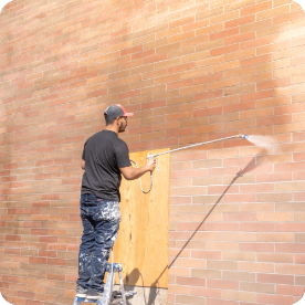 A person on a ladder pressure washes a large brick wall, preparing it for commercial painting services. Using a high-pressure hose, they clean the surface, creating a noticeable difference between the cleaned and uncleaned areas. Dressed in a black shirt, blue paint-stained pants, and a cap, they work diligently.