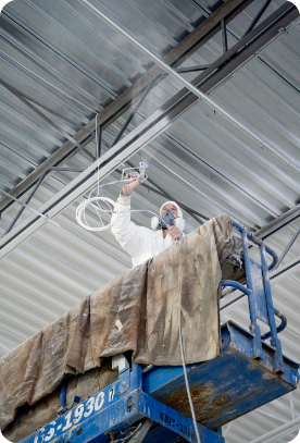A worker wearing a protective suit and mask sprays paint onto a metal ceiling using a spray gun, showcasing the precision of commercial painting services. The worker is standing on a blue scissor lift covered with a canvas drop cloth in an industrial setting with visible beams and metal panels.