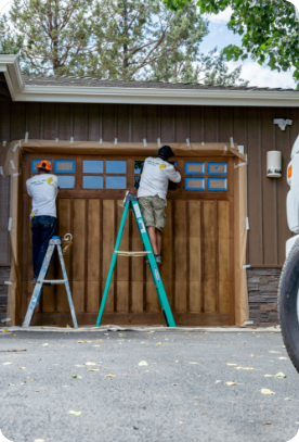 Two workers wearing white shirts and shorts stand on ladders, providing exterior painting services on a wooden garage door. The garage is part of a brown-colored house with brick accents. Trees and a partially cloudy sky are visible in the background.