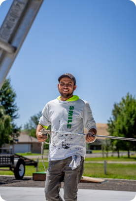A man in a white, paint-splattered shirt and cap is smiling while holding a measuring tape outdoors. The sky is clear and blue, and there are trees and a house in the background. He appears to be offering exterior or interior painting services at a construction site.