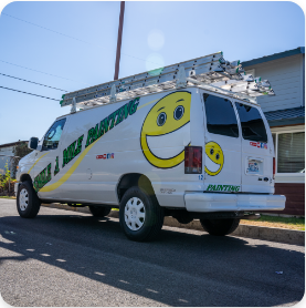 A white painting service van is parked on the street, featuring a large smiley face logo and the text "Smile & Shine Painting" in green letters on the side and rear. The van, offering both exterior and interior painting services, has ladders secured on top and is parked near a house.
