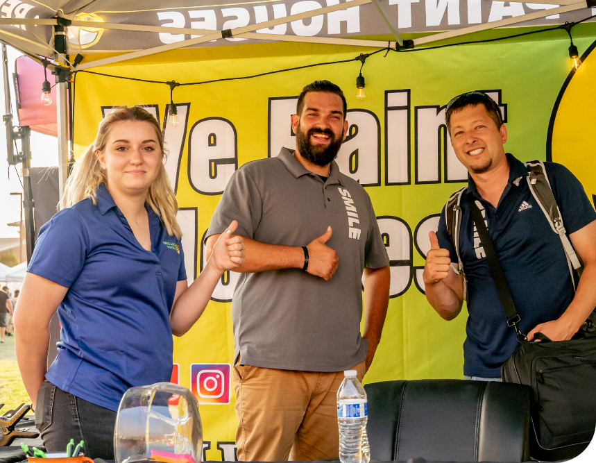 Three people stand in front of a booth under a tent with a yellow banner that reads "We Paint Houses." They all give thumbs up. The woman on the left, with blonde hair, wears a blue shirt; the bearded man in the middle wears a gray shirt, and the man on the right wears a blue shirt and backpack. A water bottle and table setup are in the foreground, advertising