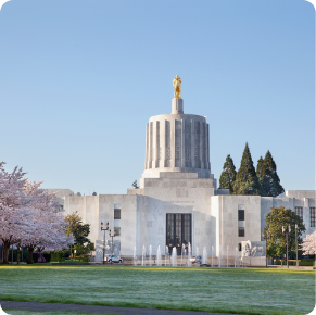 The image shows the exterior of the Oregon State Capitol building in Salem, Oregon, featuring a distinctive cylindrical dome topped with a gold statue. Cherry blossom trees and a fountain are in the foreground on a clear, sunny day.