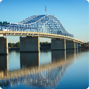 A steel arch bridge with a blue framework and white supports spans over a calm river, reflecting its structure in the water. The sky is clear and blue, with trees visible in the background under the bridge. An American flag flies from the top of the bridge, showcasing its well-maintained exterior.