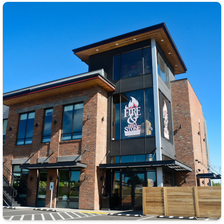 A modern brick and glass building with a large "Fire & Stone" logo displayed prominently on a corner window. The structure, boasting expert commercial painting services, features a mix of materials and has large windows, a sloping roof, and an outdoor seating area enclosed by a wooden fence.