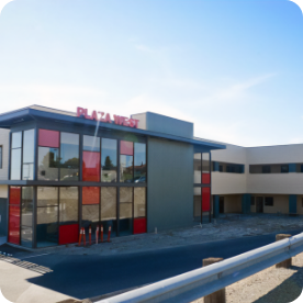 A modern two-story building with a sign reading "Plaza West" in red letters on the top. The building features large glass windows and red accents. The surrounding area appears under construction with an unpaved ground and construction cones, likely preparing for commercial painting services.