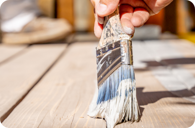 A close-up of a hand holding a paintbrush, applying white paint to a wooden surface. The background is blurred, focusing the attention on the brush and the area being painted—perfect for showcasing interior commercial painting services.