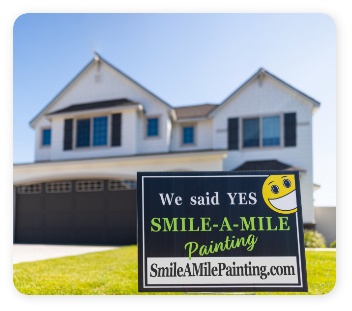 A two-story house with white siding and dark shutters stands in the background. In the front yard is a sign that reads, "We said YES SMILE-A-MILE Painting SmileAMilePainting.com," featuring a smiley face in the top right corner, proudly showcasing their exterior and commercial painting services.