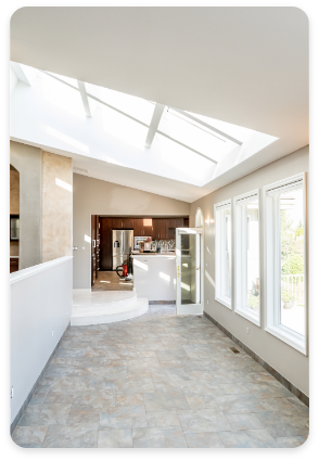 A bright kitchen and dining area featuring a skylight and large windows that fill the space with natural light. The room has tiled floors, a partially open layout, and a door that opens to an inviting patio or outdoor area. Stainless steel appliances are visible in the distance, showcasing immaculate interior design.