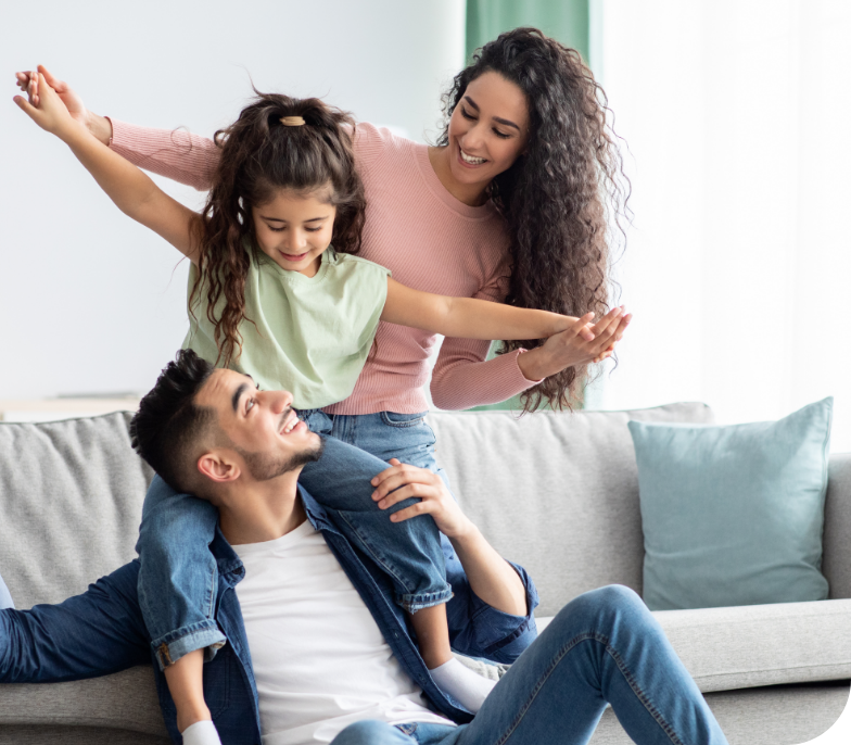 A happy family is playing together in a beautifully painted living room. The father is seated on a couch, holding his daughter on his shoulders with walls that showcase the quality of professional interior painting services. The daughter has her arms outstretched like an airplane, while the mother gently holds her hands, smiling down at them both.