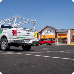 A parking lot with a white pickup truck, equipped with a ladder rack, displays a painting service logo and contact number for exterior and interior projects. In the background, there's a red truck parked and a store with an "ULTA" sign on the building. The sky is clear and sunny.