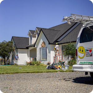 Workers painting the exterior of a white house with a black roof. One worker stands on a ladder, another on the ground. A white van with a smiley face logo and "We Love Our Cities" sticker is parked nearby. Lawn equipment and paint cans are scattered in the yard, showcasing their commercial painting services.