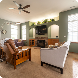A cozy living room with sage green walls, beige carpeting, and large windows. Furniture includes a leather recliner, a gray couch, and a purple chair. A flatscreen TV is mounted above a stone fireplace, with built-in shelves and cabinets on either side. Perfectly done by top-notch interior painting services.
