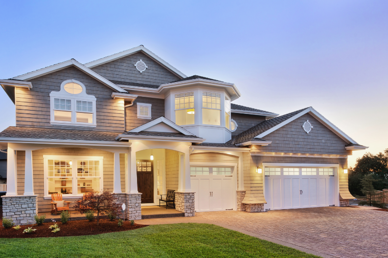 A large two-story house with grey siding and white trim, featuring multiple gables and large windows. The property includes a manicured lawn and a paved driveway leading to two garage doors. The house is well-lit, showcasing expert exterior painting services, and features a cozy front porch.
