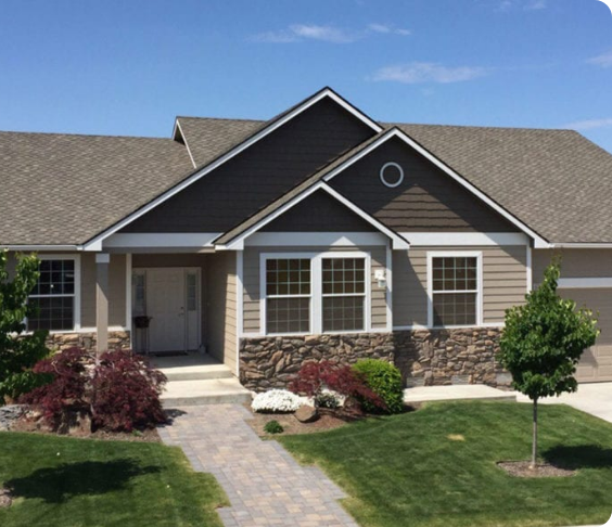 A one-story house with brown siding and stone accents is shown. It has a neatly manicured lawn, a stone walkway, and small trees and shrubs in the front yard. The sky is clear and blue, highlighting the home's pristine exterior thanks to professional painting services.