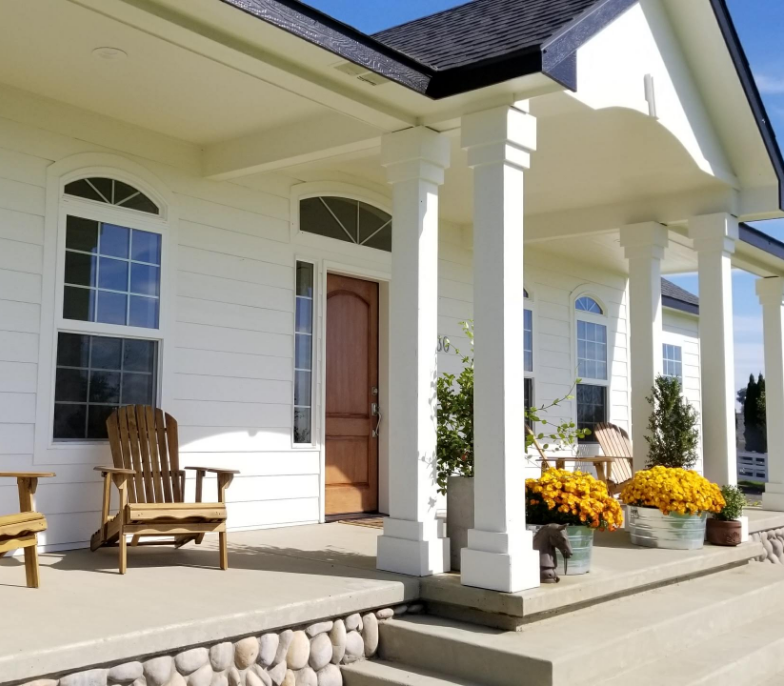 A white house with a wooden front door and a charming porch. The porch features two wooden chairs and several potted yellow flowers, adding a touch of color. The exterior is adorned with tall white columns and surrounded by a clear blue sky. Rocks line the base of the steps, enhancing its curb appeal.
