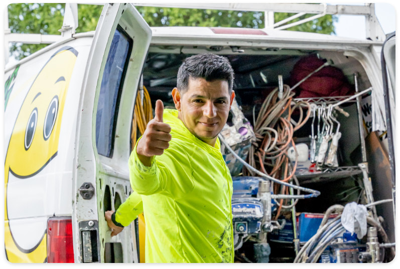 A man wearing a bright yellow long-sleeve shirt gives a thumbs-up while standing at the back of a work van. The van, open and filled with various tools and equipment for commercial painting services, has a large smiling face graphic on the rear door. Trees are visible in the background.