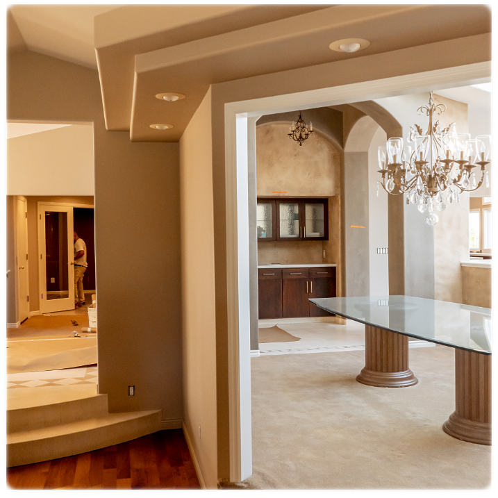 A modern, open-concept home interior featuring a partially enclosed dining area with a large glass table and an elegant chandelier. The background shows a kitchen with dark wood cabinets. A worker is visible in a hallway, likely providing commercial painting services, indicating ongoing construction or renovation.