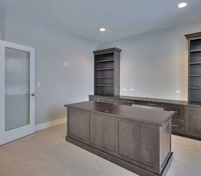 A modern, unfurnished office space featuring a wooden desk in the center. On either side of the desk are built-in dark wood bookshelves and cabinets against light-colored walls. The door, partially open, has frosted glass panels. The floor is carpeted, ideal for interior commercial painting services.