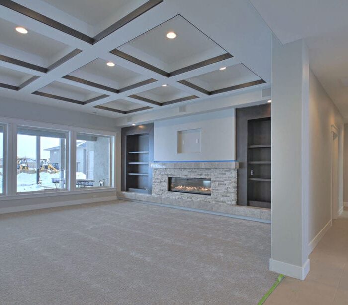 A modern living room with beige carpet flooring and a recessed ceiling with geometric patterns. Large windows provide natural light and a view of a snowy landscape. The focal point is a sleek fireplace embedded in a wall with built-in shelves on either side, showcasing impeccable interior design.